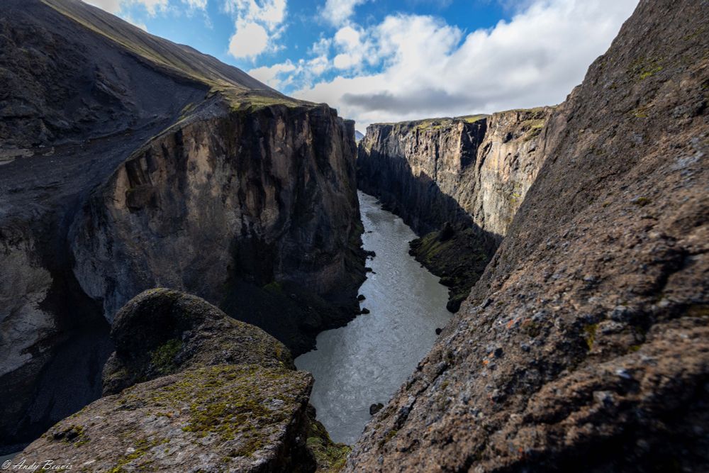 A narrow and deep canyon is in the centre of the view, the canyon curving slightly left into the distance.  The canyon sides are solid rock. The river in the bottom is a milky in colour. The sky is blue with some fluffy white clouds.