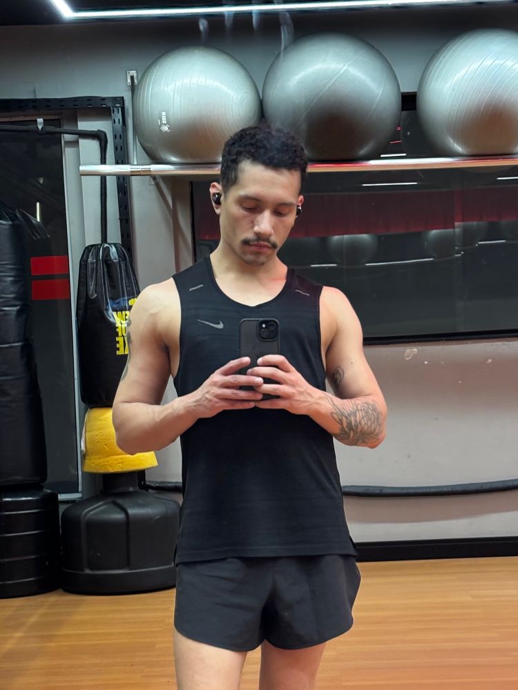 uma foto minha no espelho da academia, estou de regata preta e short preto segurando o celular com duas mãos em frente ao corpo. meu ombro está um bafo 