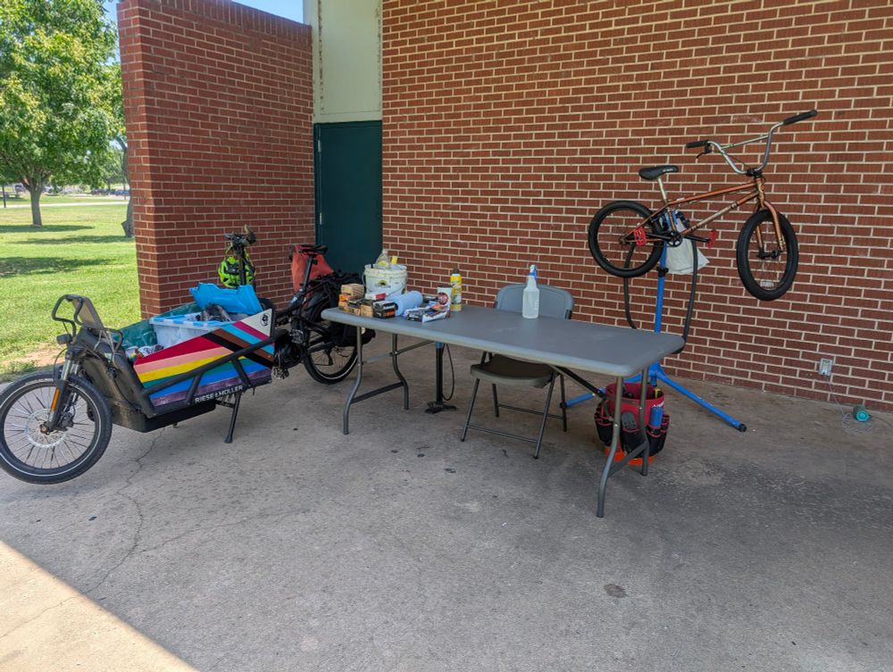Along the side of a brick park building, a cargo bike with an Ally Flag painted on it is parked next to a table and bike stand. A BMX bike is on the bike stand, and spare tubes and bike cleaning supplies are on the table.