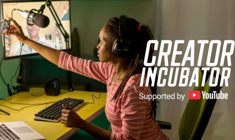A person wears headphones and sits in front of a large screen with a keyboard. They reach forward, taking a selfie 