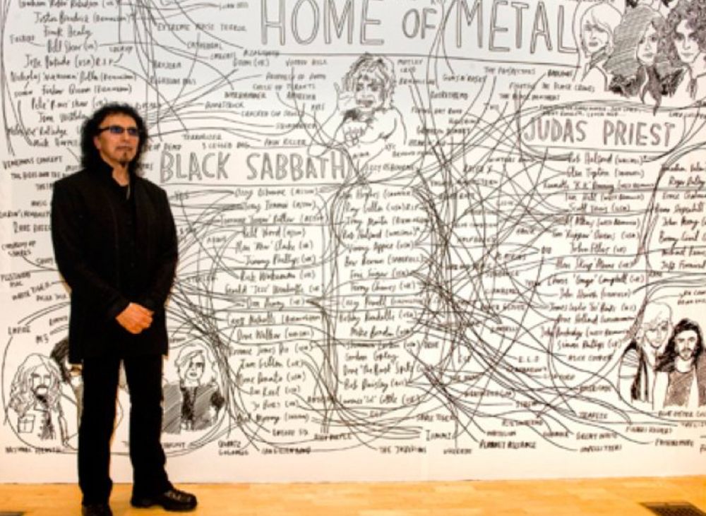 Toni Iommi stands in front of a white board featuring tributes and drawings of Black Sabbath
