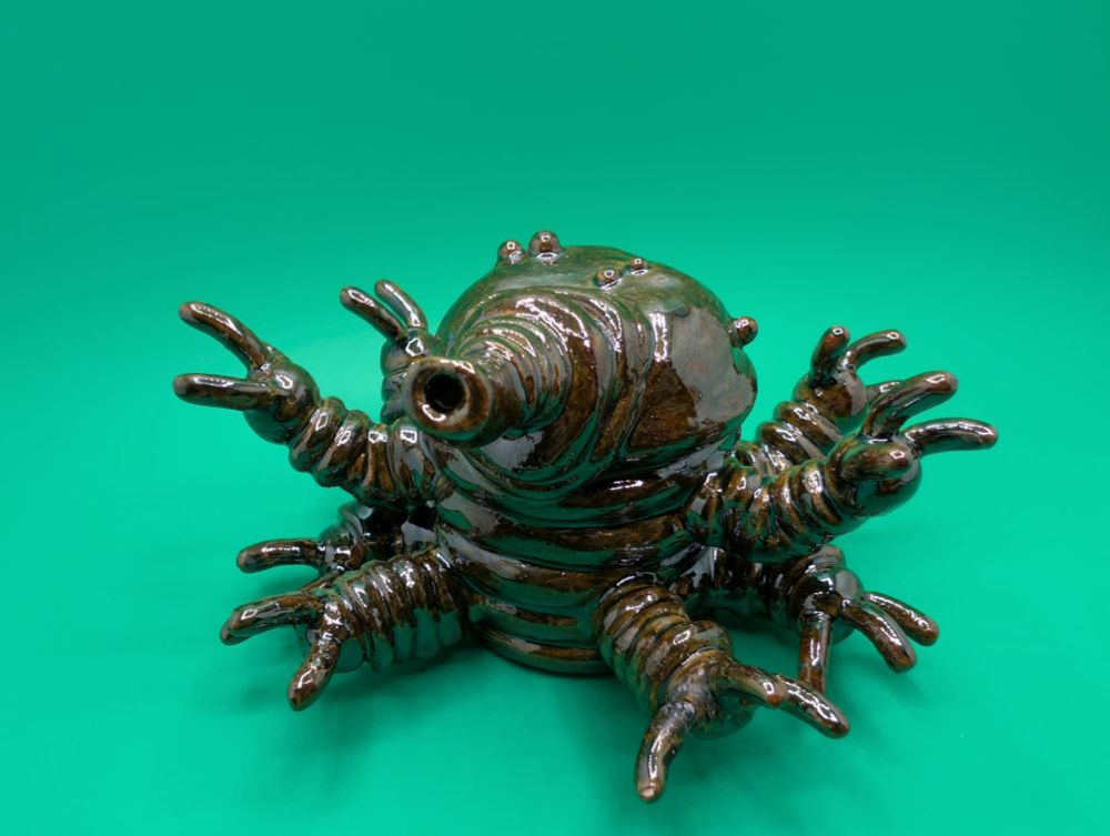 A  view from the right of a stoneware ceramic sculpture of an eight armed creature with a long proboscis.  The sculpture has a very shiny glaze with deep browns and greens.  The background is light green.