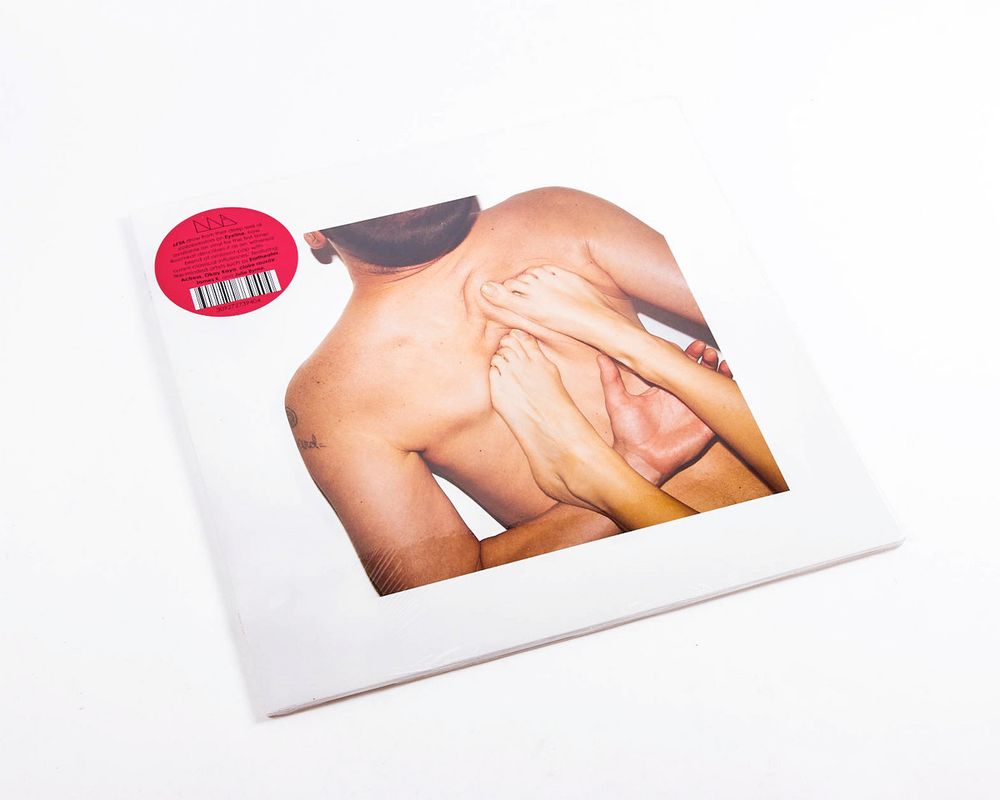  A vinyl record with a minimalist white sleeve featuring a provocative photograph on the cover: a shirtless person seen from behind is entwined with several bare arms and legs, creating a tangled and intimate composition. A circular red marketing sticker with a barcode is affixed in the top left corner.