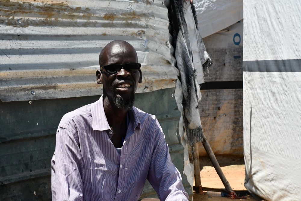  David Liep, a South Sudanese refugee returnee with a visual impairment, at Mangaten camp in Juba, South Sudan. David is wearing a buttoned, light blue shirt and dark glasses. He is sat in front of a temporary structure in Mangaten, made from metal sheets and plastic tarps.