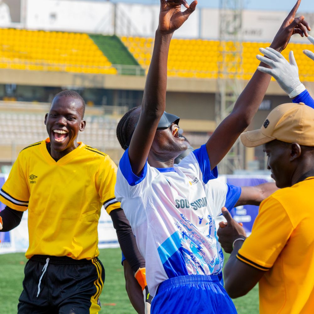 South Sudan Blind Football player in white and blue lifts arms while interacting with team staff on the pitch.

