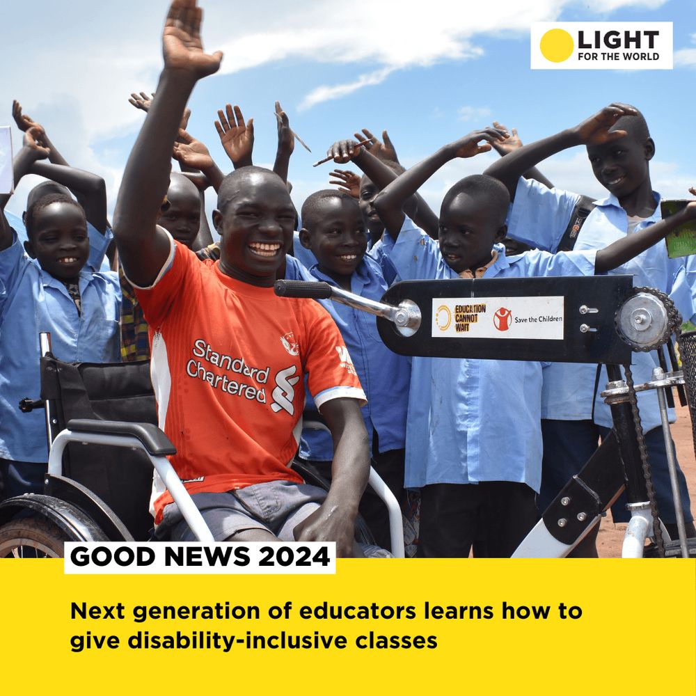 A man in a wheelchair is waving towards the camera. Behind him, a group of children wearing blue shirts is imitating his movements and smiling. 