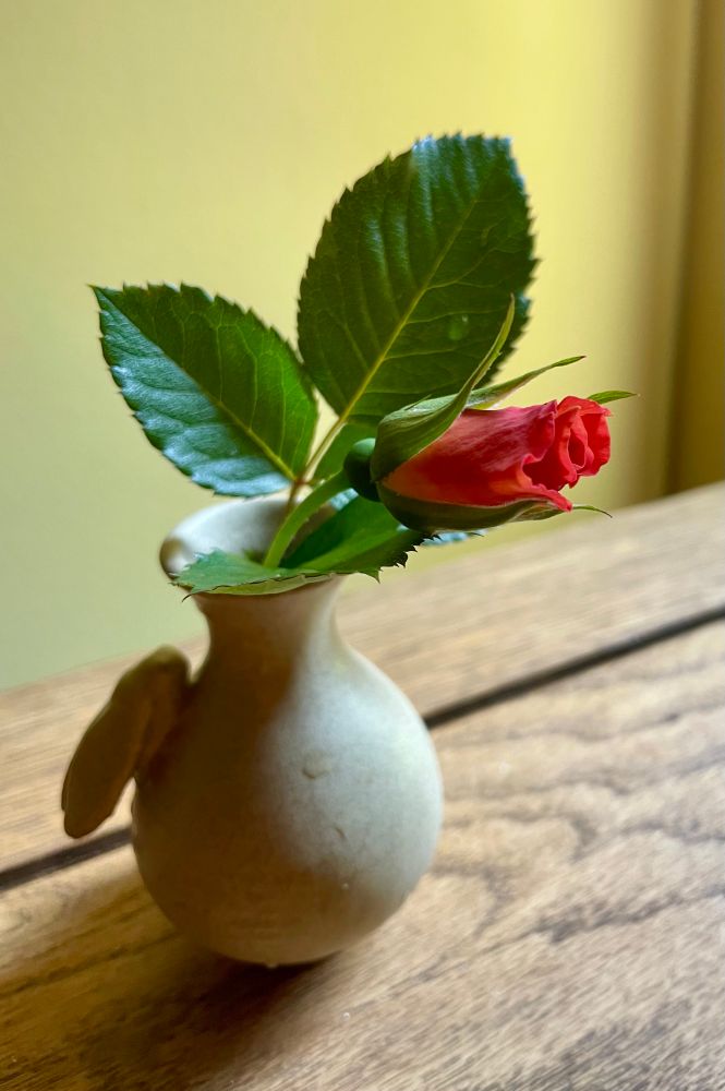A tiny orange rosebud in a tiny vase on a wooden table 