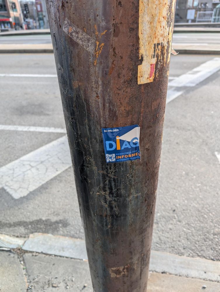 Photo of a utility pole in a city intersection, with a square blue sticker attached to it. The sticker reads:

DI-AG.ORG

DEMOCRATS FOR AN INFORMED APPROACH TO GENDER

The logo shows the letters "DIAG" in light blue, with the I written to look like a tall orange lighthouse shining out a light pointing to the right.