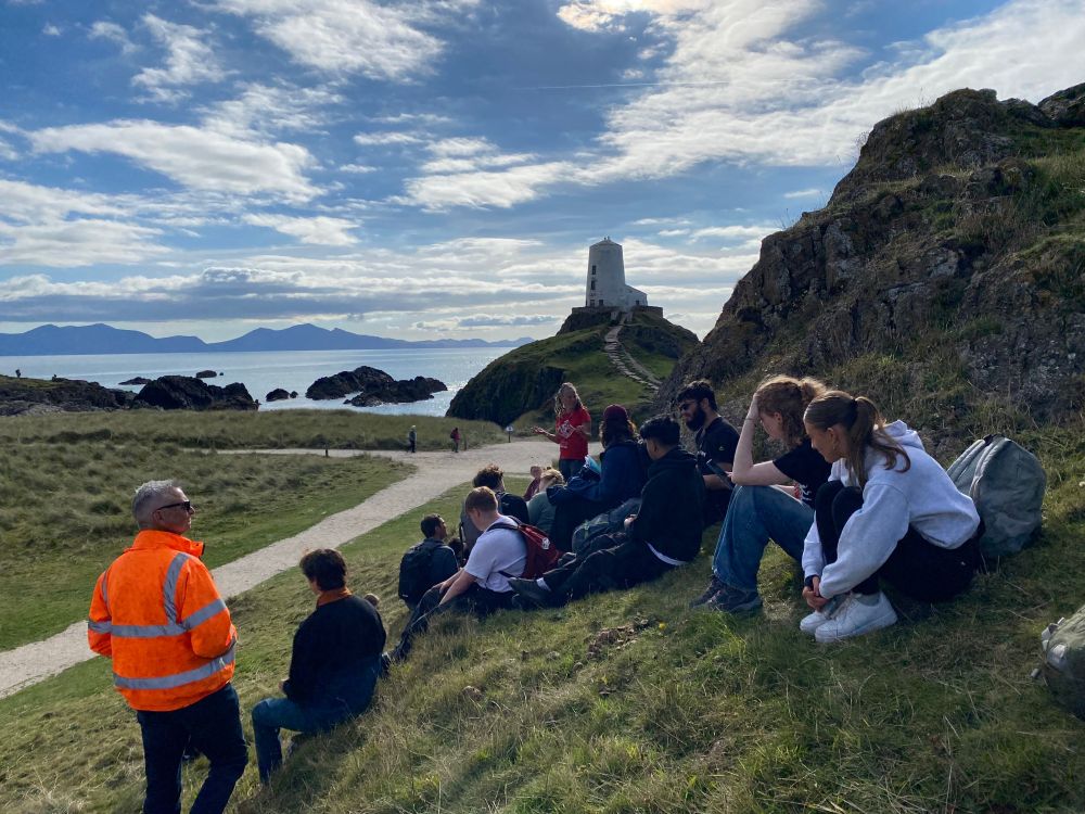 Students and lecturers sitting on the grass with a lighthouse beyond
