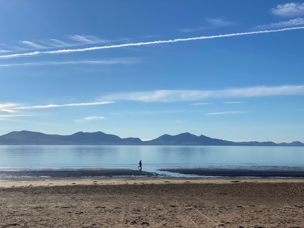A lone walker on a beach with very calm sea and mountains in the background
