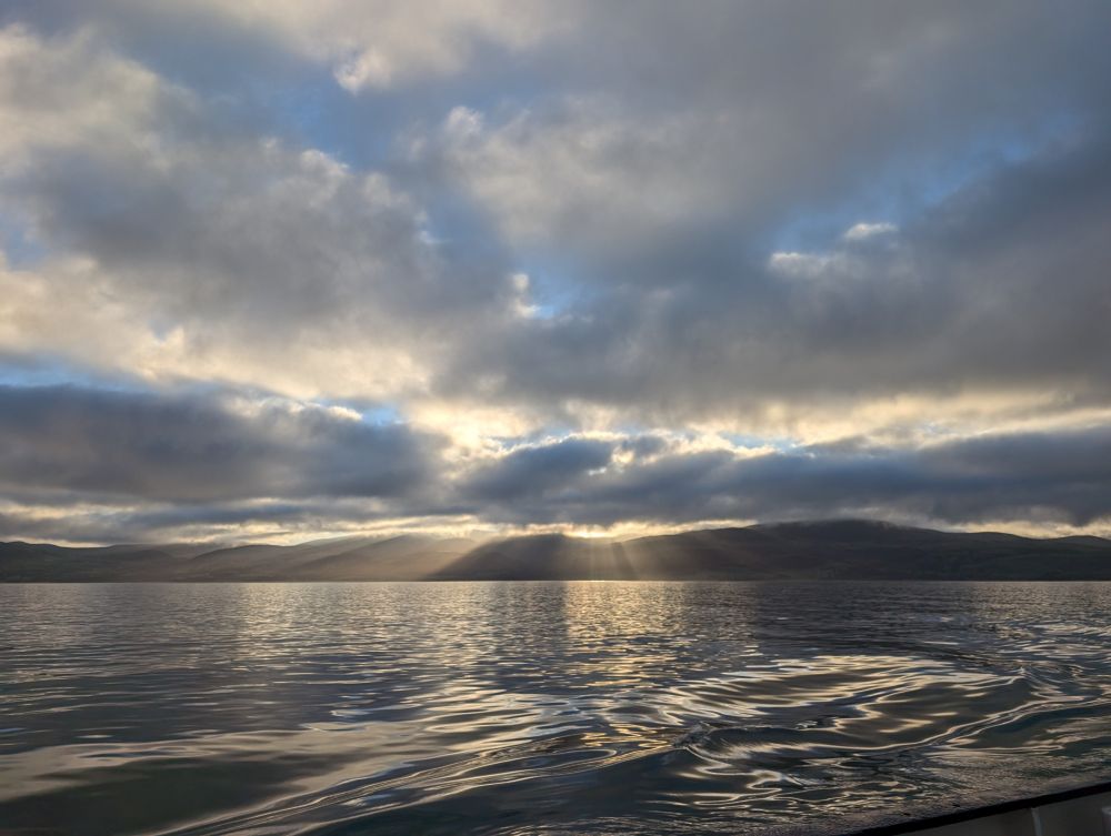 Sun beams shining through clouds over the mountains beyond a glassy sea