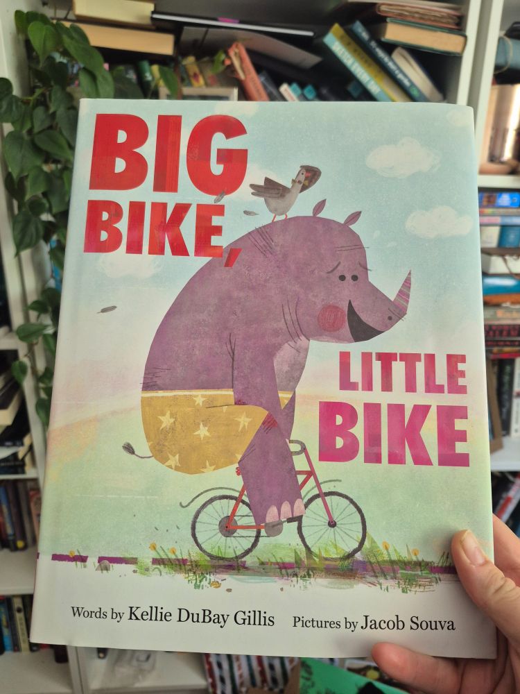 A picture book with the title Big Bike, Little Bike, Words by Kellie DuBay Gillis, Pictures by Jacob Souva. An illustration of a purple rhinoceros in yellow shorts rides a bike, a bird sits on his head