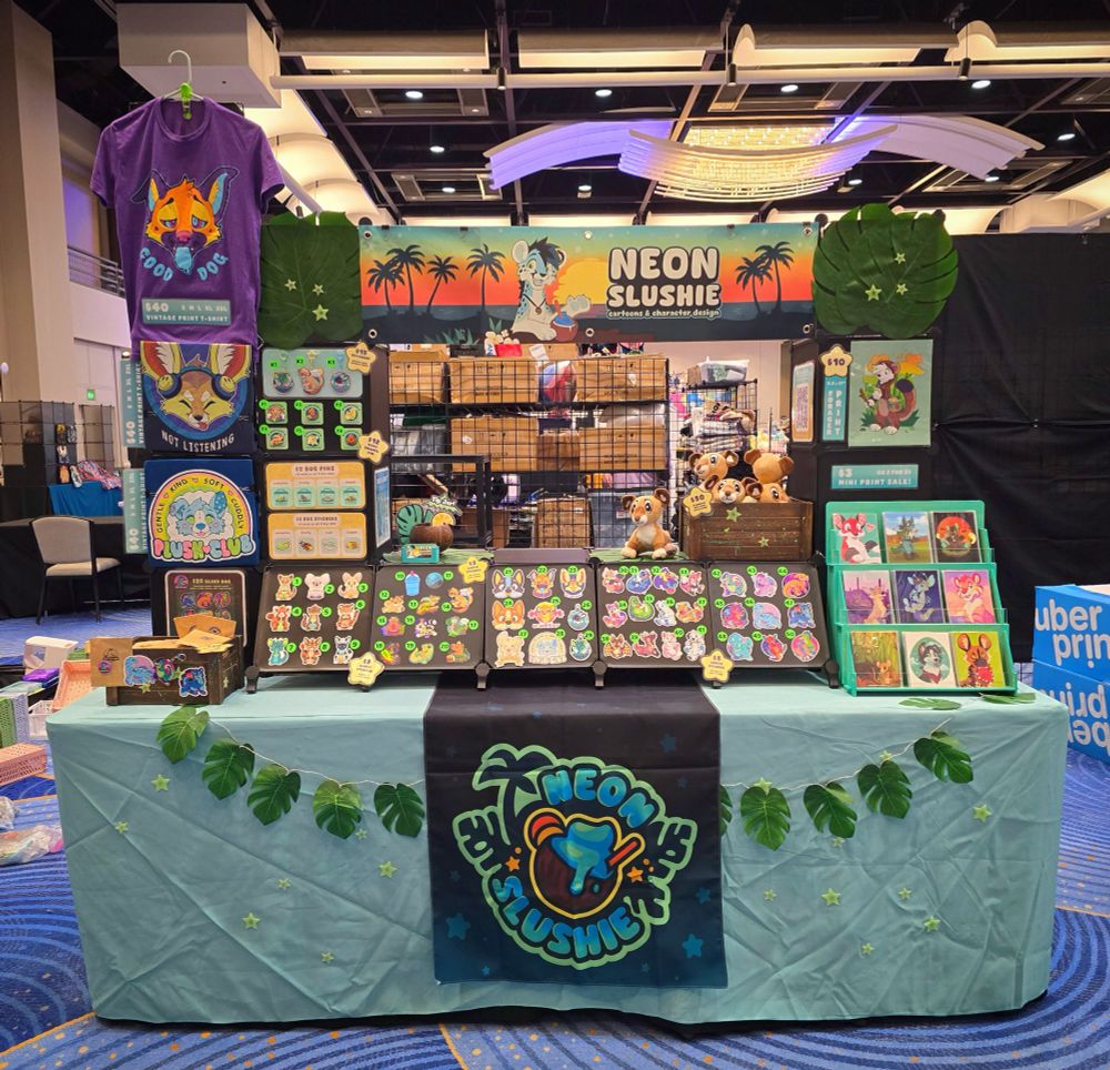 A photo of my art booth at Furry Weekend Atlanta. Furry T-shirts, stickers, enamel pins, keychain charms, prints, and pride thylacine plushies! 🎨 #FWA #FurryWeekendAtlanta #FurSale #FurryArt