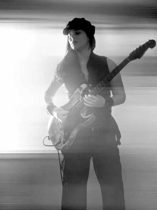 A high key black and white photo of Kiri Bereznai of the post punk rock band, The Rapid Onset. Kiri is dressed in black, awash with rays of light, playing a custom Squier Strat with a heavily distressed, naturally achieved relic finish. 