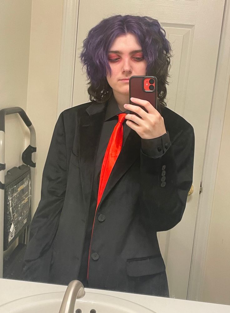 Mirror selfie of my Revenge era Gerard Way costume. I’m looking down at my phone in the picture. I’m wearing a black button up, a black velvet suit jacket, a red tie, and black corduroy pants (not pictured). I’m also wearing a mix of red and brown eyeshadow 