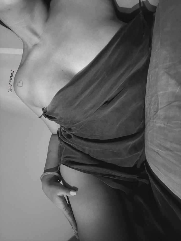 Melody in monochrome, wearing a black, satin slip dress. The strap is dangerously low, showcasing her cleavage. She's laying on a bed, with her thigh slightly propped up. She has one hand on her upper thigh.