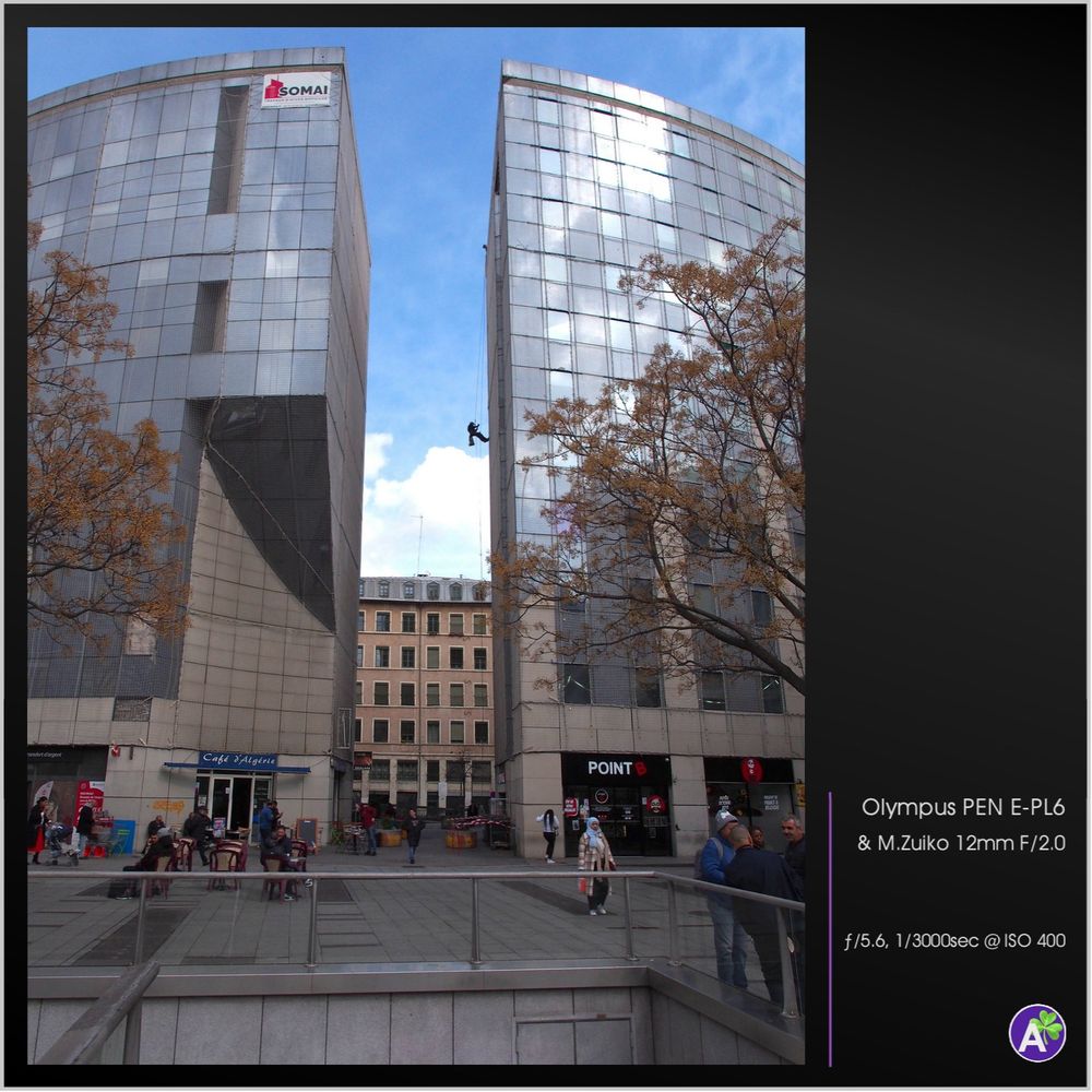 a color outdoor photograph. a window washer rappels down the side of a building between two matching glass buildings. the two buildings each form a quarter of a semi-circle. many people mill about on the sidewalk and in the alley between two buildings. there is a café with some outdoor tables. in the foreground are stairs descending as if to a parking garage or subway station.