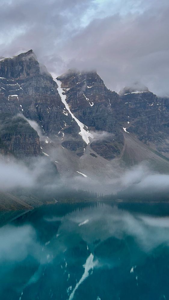 The snowy mountain peaks stand tall above Moraine Lake's crystal blue water as numerous cloud banks brush past them.