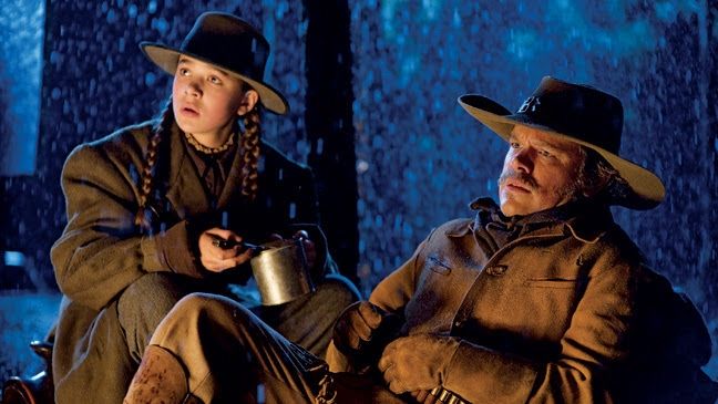 Hailee Steinfeld as the teenage protagonist Mattie Ross (with Matt Damon) in the Coen brothers’ "True Grit." She wears an oversize coat and hat, hair in two braids, and holds a pot of food for Damon's character, who's injured. The warm light of a campfire illuminates them as snow falls in the background. 