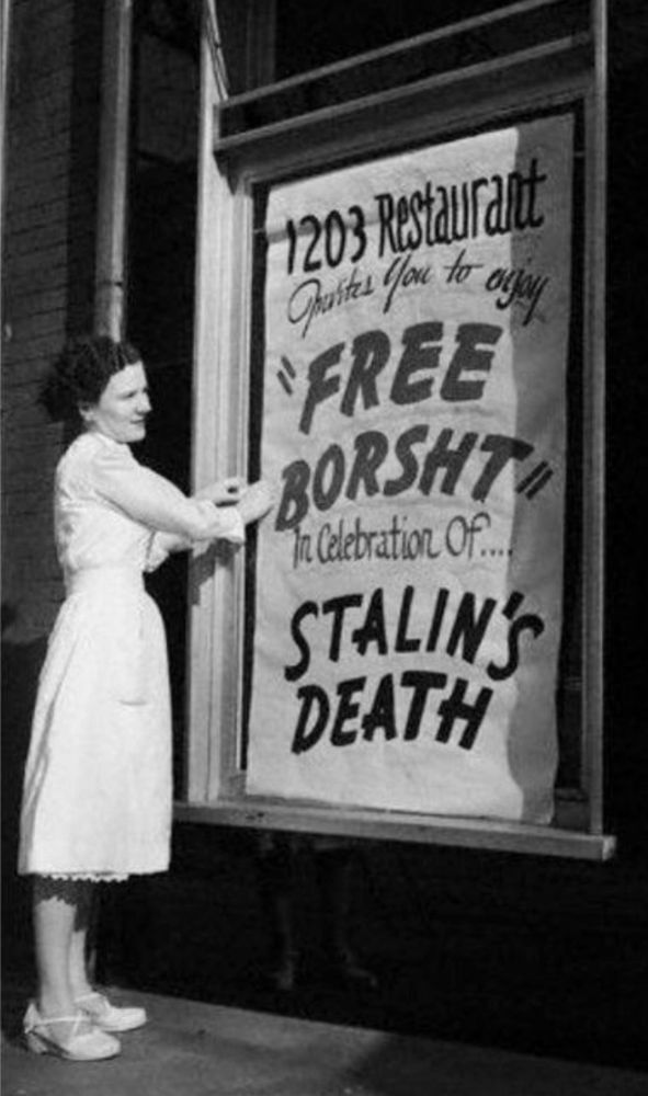 Old black & white photo of a woman standing by a large poster outside a restaurant that reads 'FREE BORSHT IN CELEBRATION OF STALIN'S DEATG'