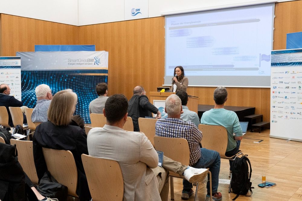 Eine Referentin spricht vor einem aufmerksamen Publikum über die Phasen der Energiewende. Hinter ihr ist eine Präsentationsfolie zu sehen. Links und rechts stehen Roll-Ups der Smart Grids-Plattform BW und des ZSW. Die Veranstaltung findet in einem modernen Konferenzraum mit Holzverkleidung statt.