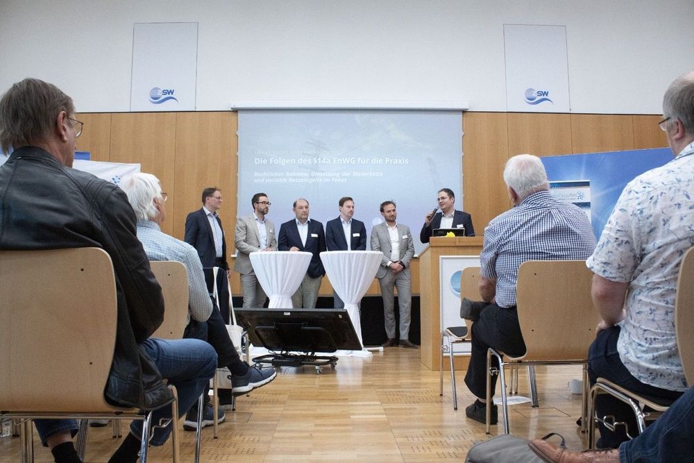 Sieben Männer stehen auf der Bühne bei der Paneldiskussion „Die Folgen des § 14a EnWG für die Praxis“. Im Fokus stehen rechtlicher Rahmen, Steuerkette und variable Netzentgelte. Die Teilnehmenden im Saal verfolgen die Diskussion aufmerksam.