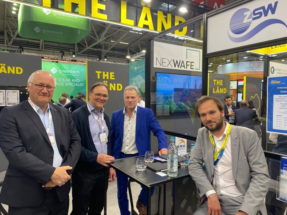 Vier Männer am Stand von ZSW und NexWafe auf der Messe The smarter E 2025 in München. Sie stehen bzw. sitzen an einem Stehtisch mit Getränken und blicken in die Kamera. Im Hintergrund ist das Branding von „THE LÄND“ sowie weitere Messestände und Besucher zu sehen.
