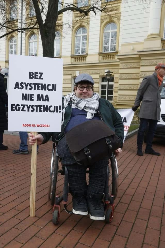 Na zdjęciu mężczyzna na wózku z transparentem w rece „bez asystencji nie ma egzystencji” na tle budynku kancelarii premiera. Mężczyzna ubrany jest w ciemne spodnie, sweter i kurtkę, arafatke, okulary oraz szary kaszkiet. 