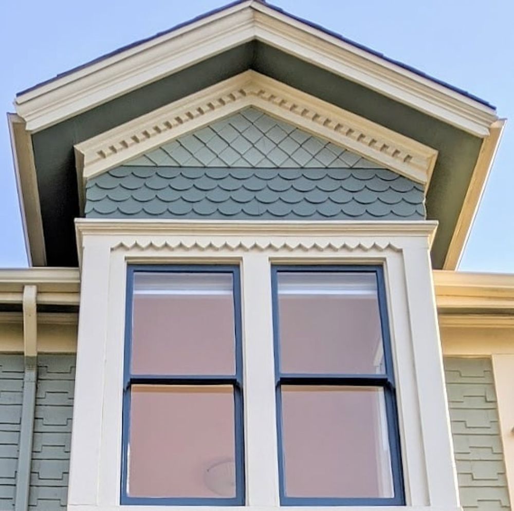 Box bay window with two blue double-hung wood windows and green and cream decorative woodwork on the surrounding house