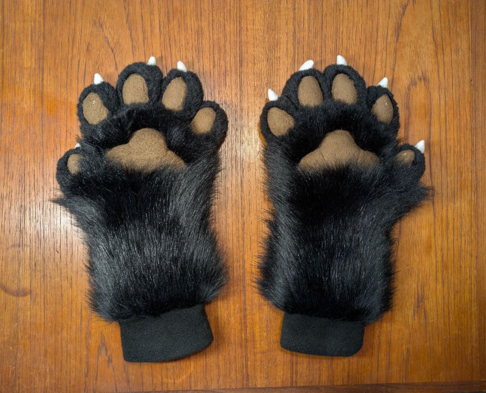 Photo of black fursuit handpaws with white claws and black pawpads on a table. Both turned palm up.