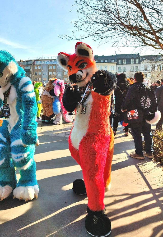 Fursuit photo of Loimu The Fox pointing his paws towards the camera in a finger-gun pose. A lot of other furs can be seen around as it's right after the NFC fursuit parade.
