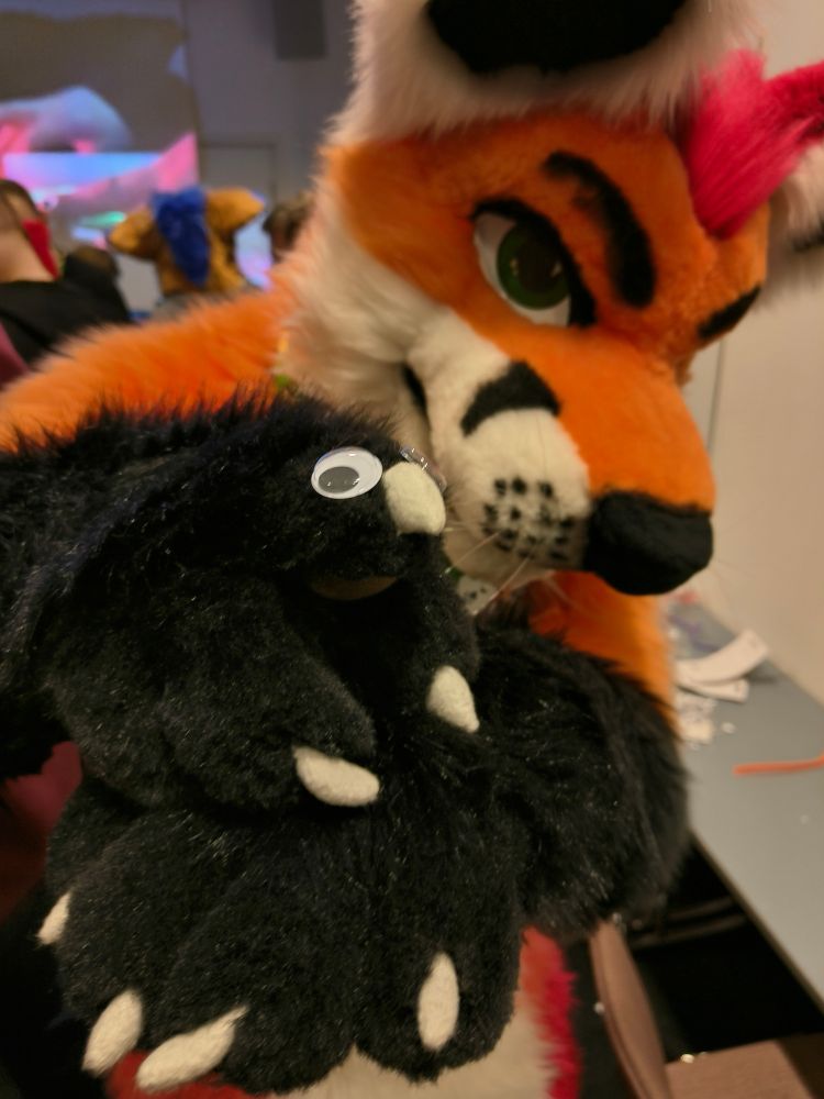 Fursuit photo of Loimu The Fox showing off his hapsiainen with him looking at the camera from the background. Hapsiainen is a small animal that looks suspiciously like Loimus handpaw with some googly eyes.
