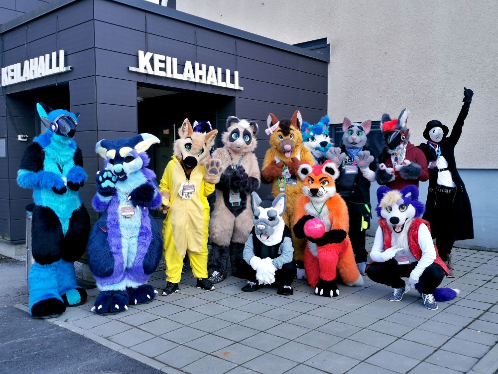 Group fursuit photo of a bunch of furries in front of the entrance to a bowling alley. It says Keilahalli, or bowling alley, on top of the entrance behind.