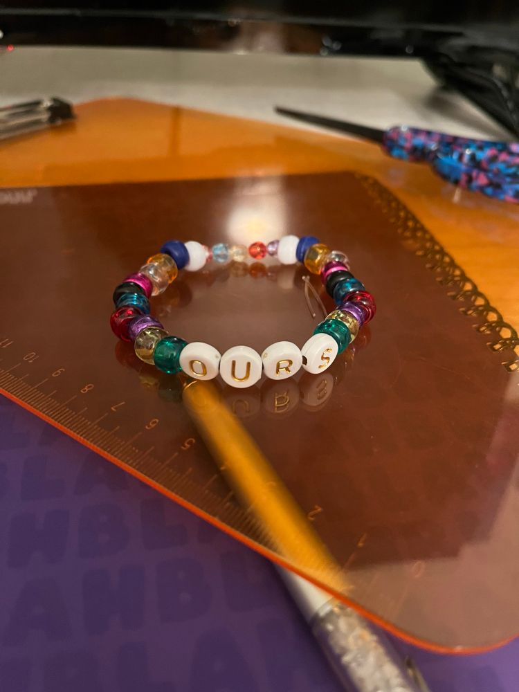 A friendship bracelet that says “Ours,” on it. It is very colorful with all the colors of the rainbow on it and then some. All the albums are represented in order.