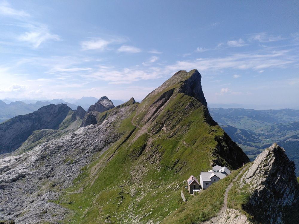 View of a rugged mountain range, karst outcrops, and green meadows from above, against a blue sky with some light clouds. The picturesque mountain inn "Tierwis" can be seen from atop. Imagine a mild breeze and the rolling calls of a chatter of Alpine Choughs.