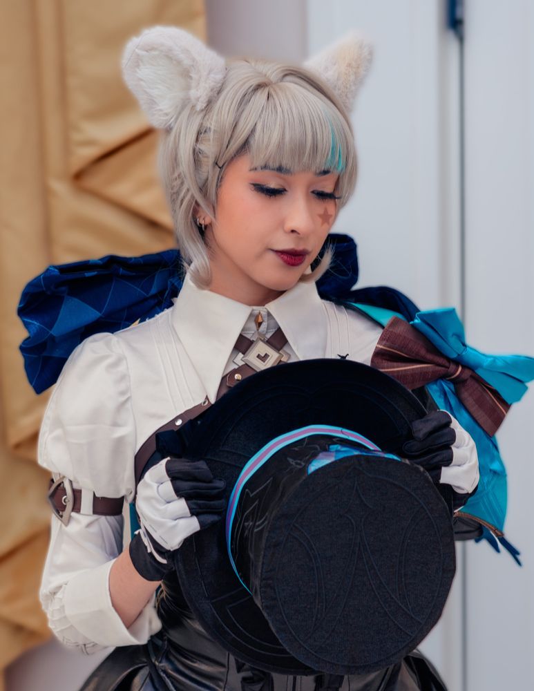 Onyx is dressed as Lynette from Genshin impact facing the camera. They’re holding a black tophat in front of their chest and looking down and too the right with their eyes shut