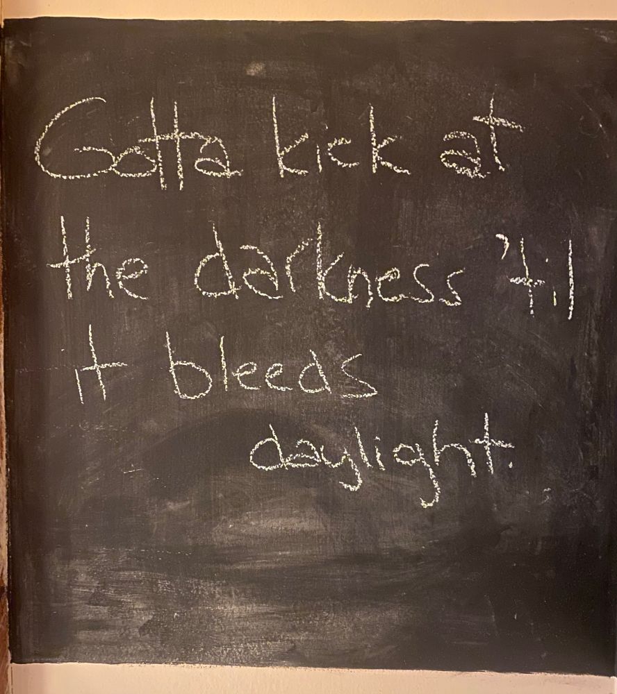 Chalkboard with the words “Gotta kick at the darkness til it bleeds daylight.”