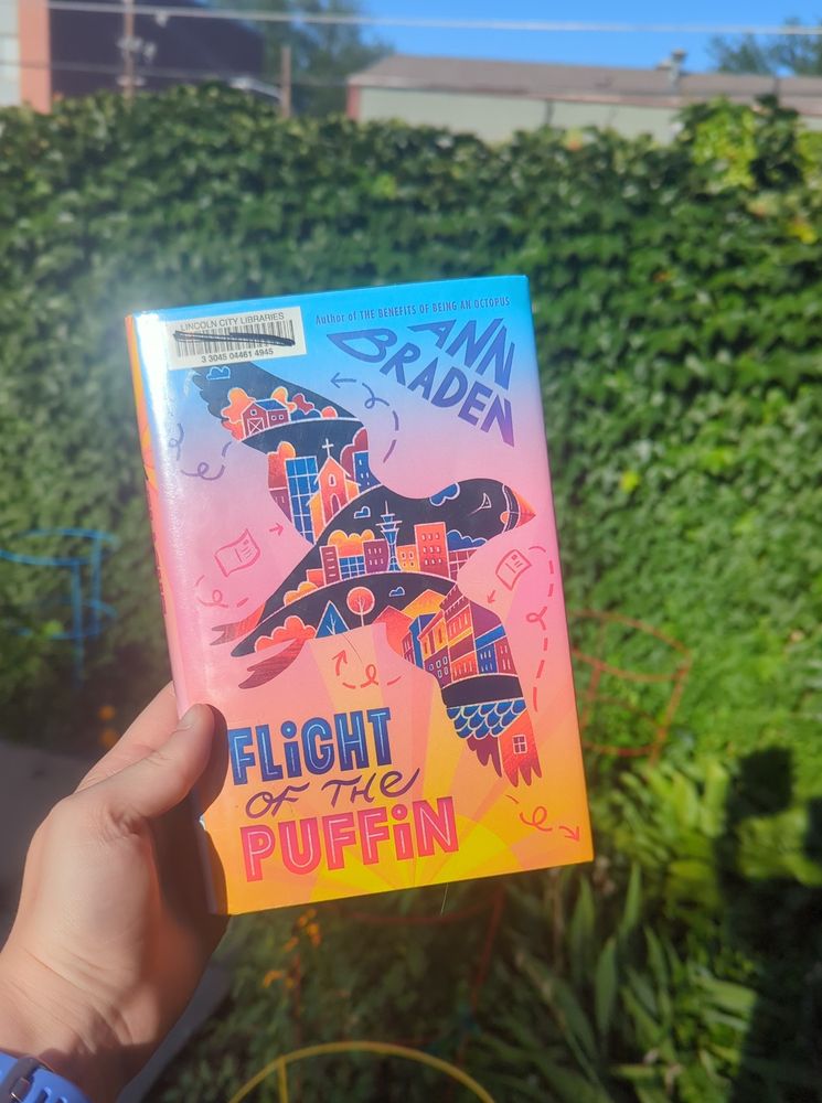 A hand holds "Flight of the Puffin," a middle-grades novel by Ann Braden, in front of an ivy-covered wall