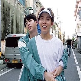 Seokjin and Hoseok in white t-shirts and teal colored jackets. Jin is hugging Hobi from behind.