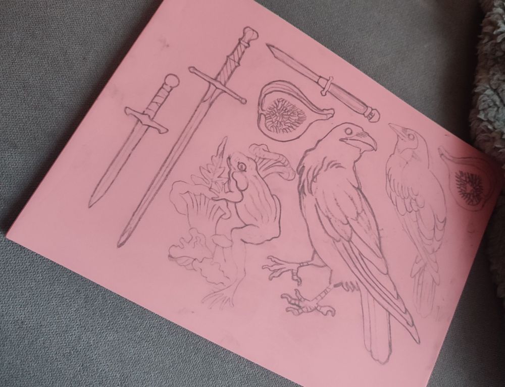 A photo of a pink linoleum bloc. On it are pencil drawings of two crows, various blades, two figs and a frog climbing mushrooms. Nothing is carved yet.