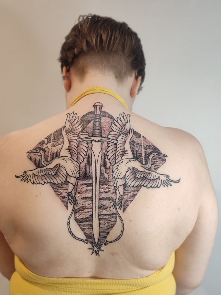 A photo showing the fresh back tattoo. Two herons are flying on either side of the spine. At the center, a sword is cutting a rope attached to their leg. The birds are flying in front of a oceanic/forest landscape.