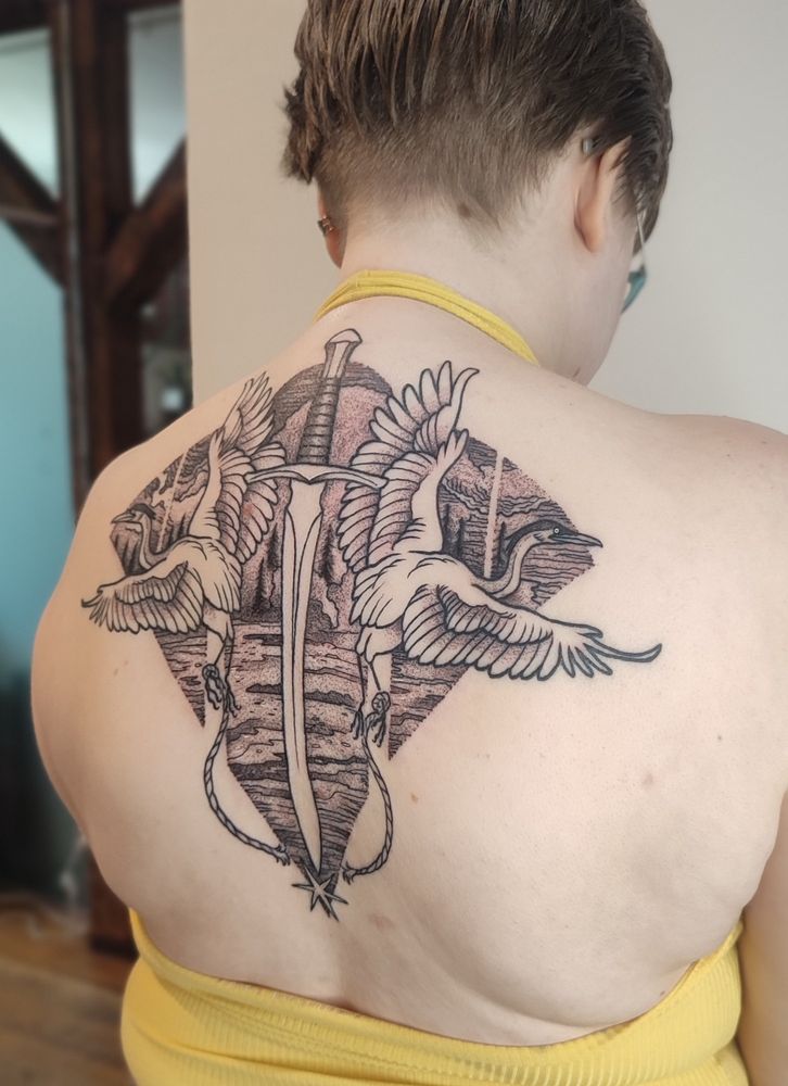 A photo showing the fresh back tattoo. Two herons are flying on either side of the spine. At the center, a sword is cutting a rope attached to their leg. The birds are flying in front of a oceanic/forest landscape.