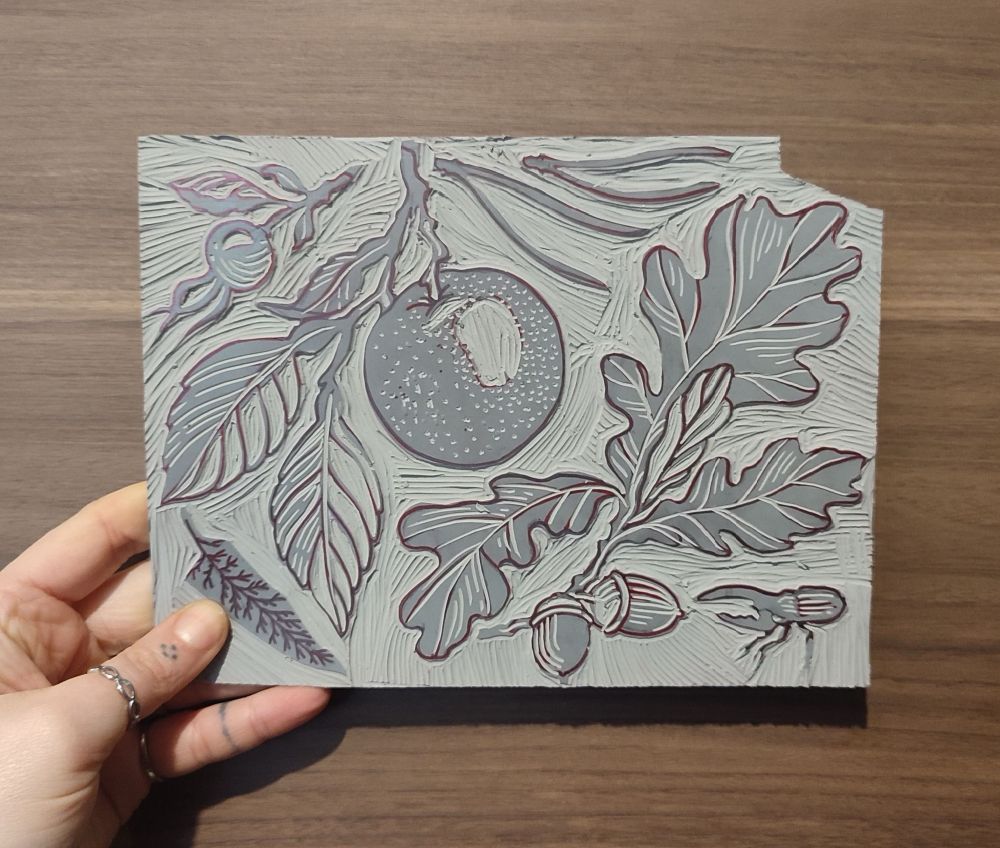 A block of linoleum, carved, with some line-work drawn onto it. The drawings represent plants: oak branch, orange fruit, pine needles, rose hips and one scarab.