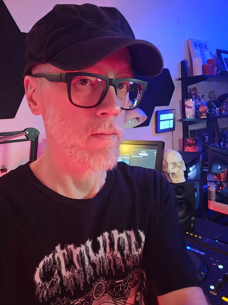 Photo of me (white, male, grayish light red beard) wearing a black cap, glasses, and t-shirt with text on it reading "Cthulhu". I'm standing in front of my DJ workstation. 