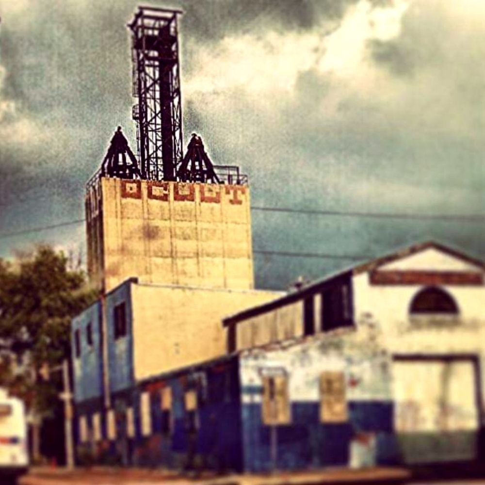 Photo of a long abandoned multistory industrial building spanning an entire city block made of brick, wood, steel, and concrete. Its highest portion has large graffiti that reads "OCULT", and has three steel girder structures on its roof. 
