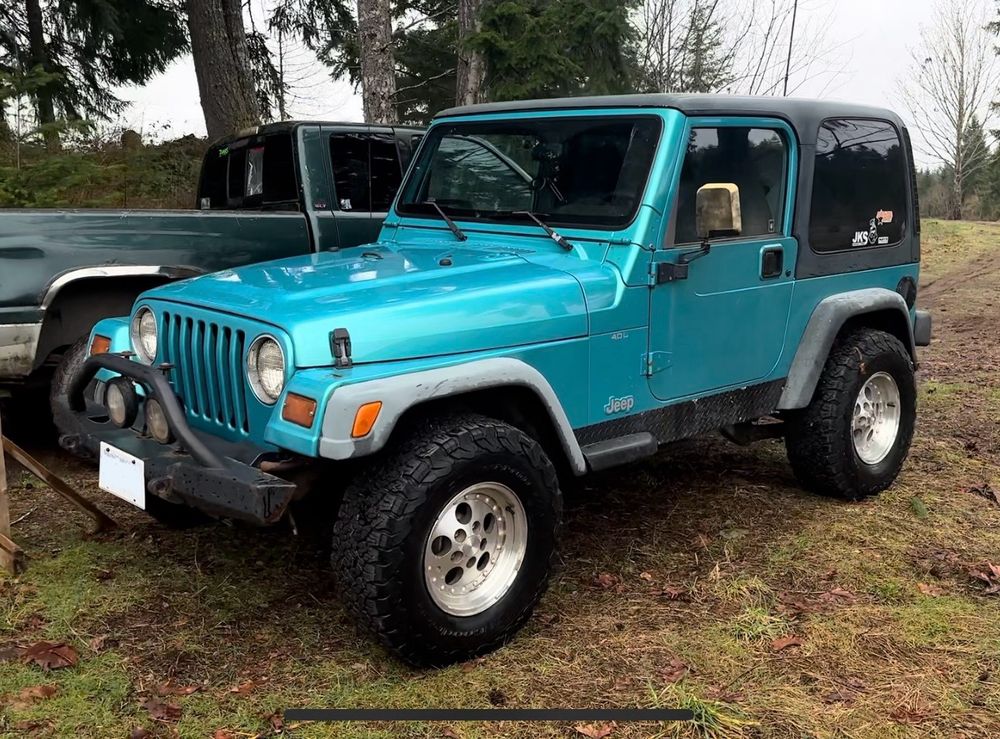 A jade satin (teal) Jeep Wrangler TJ sitting on a dirt path with a green pickup truck and trees behind. 