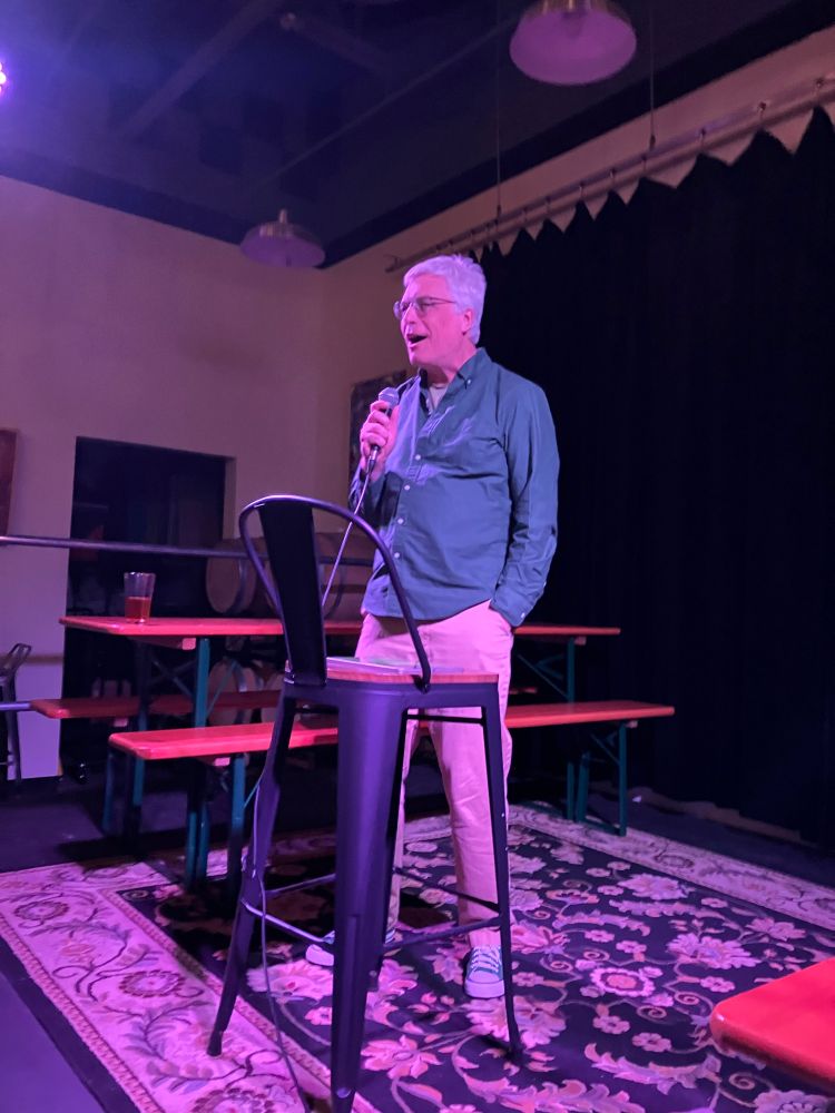 David standing on stage under a purplish light, one hand holding a mic and the other in his pocket, looking happily engaged in telling his story with his eyes on the audience.