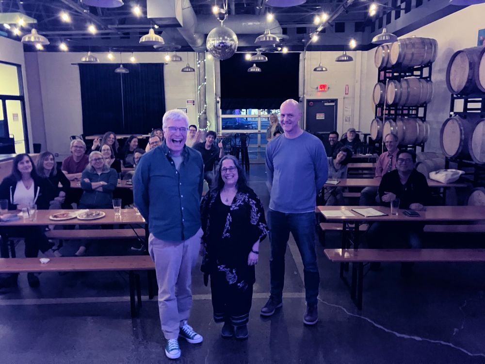 Three fantastic writers standing in front of several wooden bar tables of attendees. There are 26 people in the room, with wooden barrels along one side.