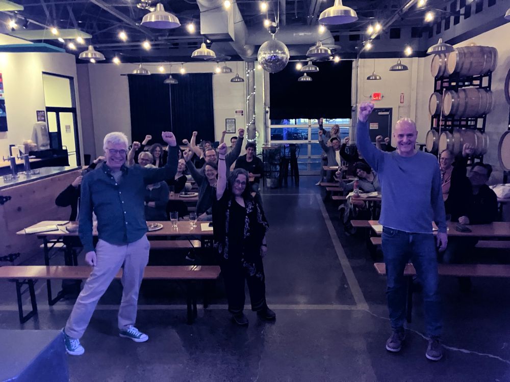 Three fantastic writers standing in front of several wooden bar tables of attendees. There are 26 people in the room, with wooden barrels along one side. In this shot, everyone’s fist is in the air.