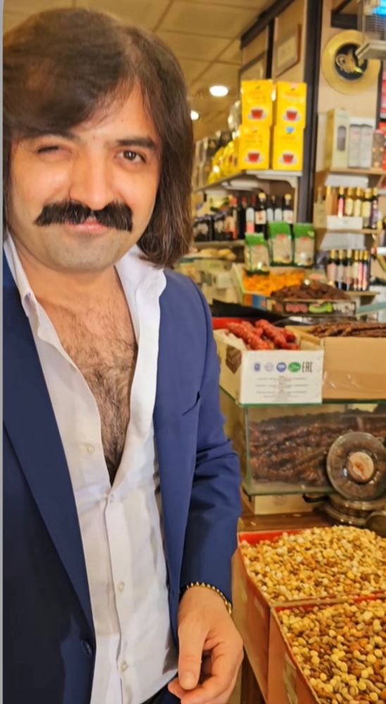 Ein zwinkernder Mann mit hammer Frisur, gepflegtem Schnäuzer und üppigem Brusthaar